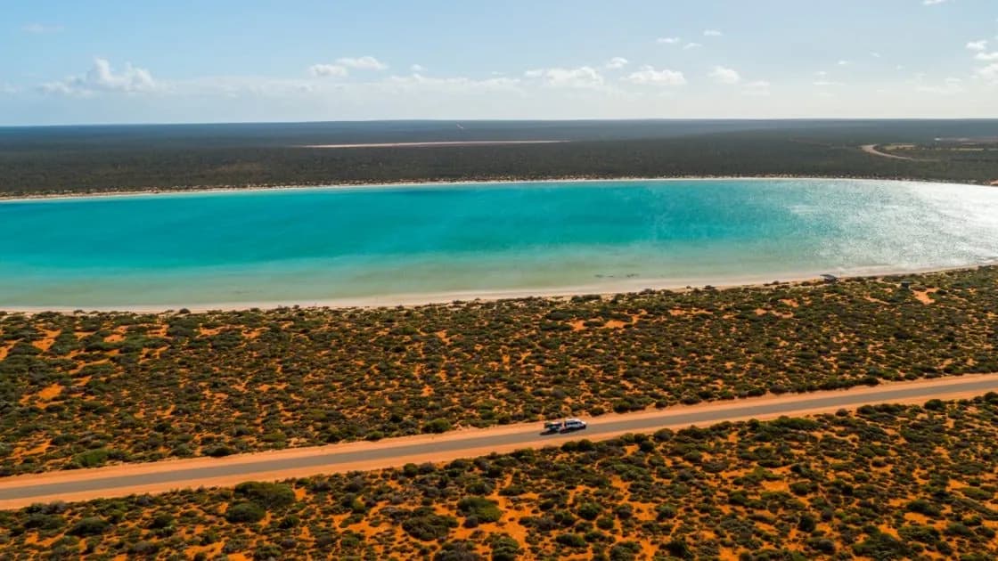 Aerial view of a sealed road through red sand and scrub beside a turquoise bay in Western Australia.
