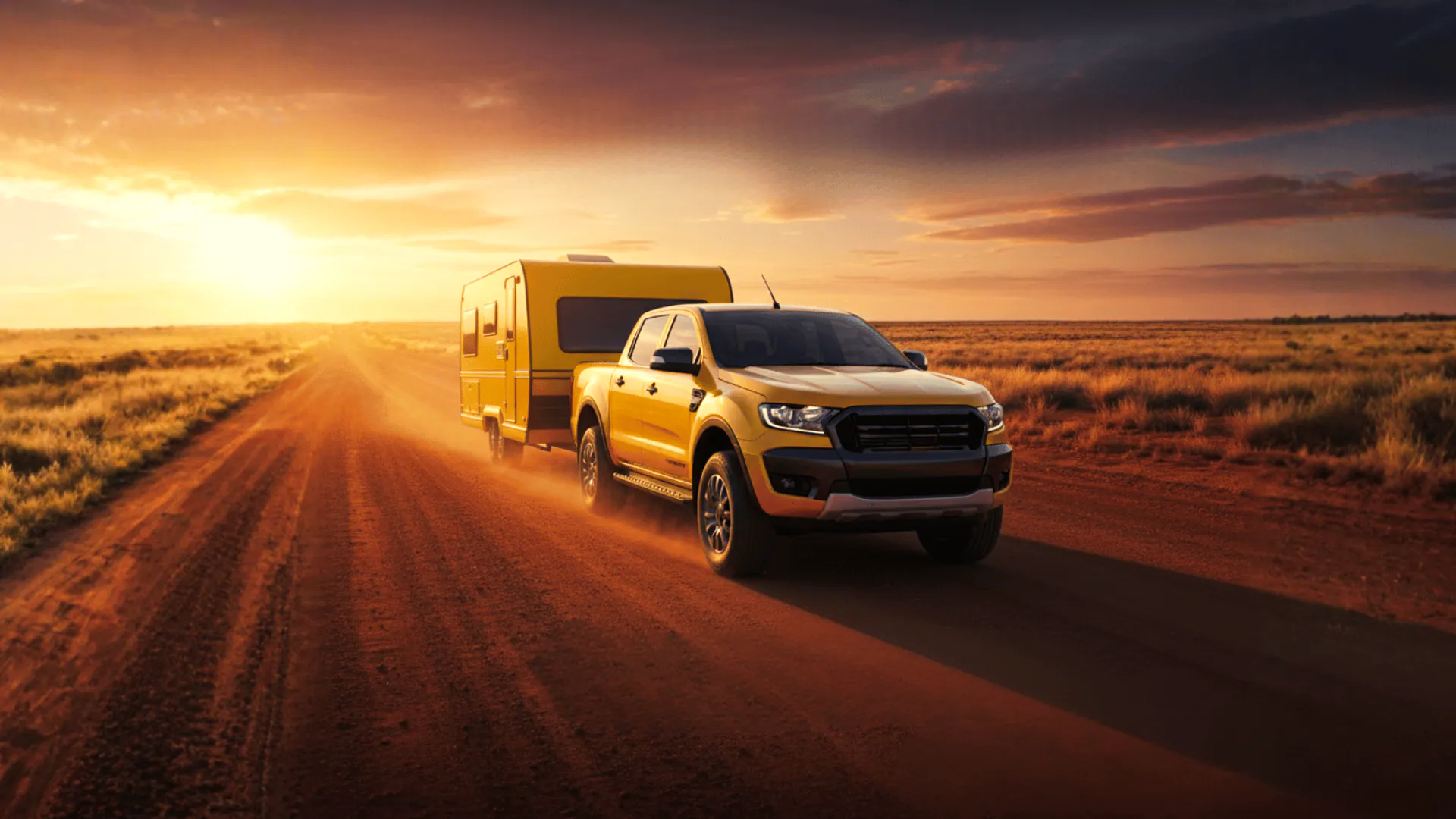A yellow ute towing a caravan on a red-dirt outback road at sunset.